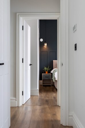 A hallway leading to a bedroom with a wooden floor, a bedside table with a plant, and a hanging light fixture. - Perfect Booking 2 Bed Flat Near Cabot Circus (Bristol)