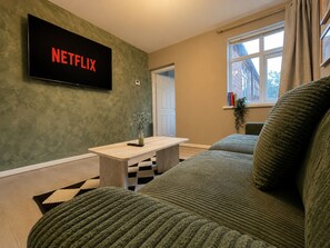 A living room with a flat-screen TV mounted on the wall displaying the Netflix logo, a wooden coffee table, a green sofa, and a window with a view of buildings outside. - Perfect Booking 2 Bed Home w Street Parking (Coventry)