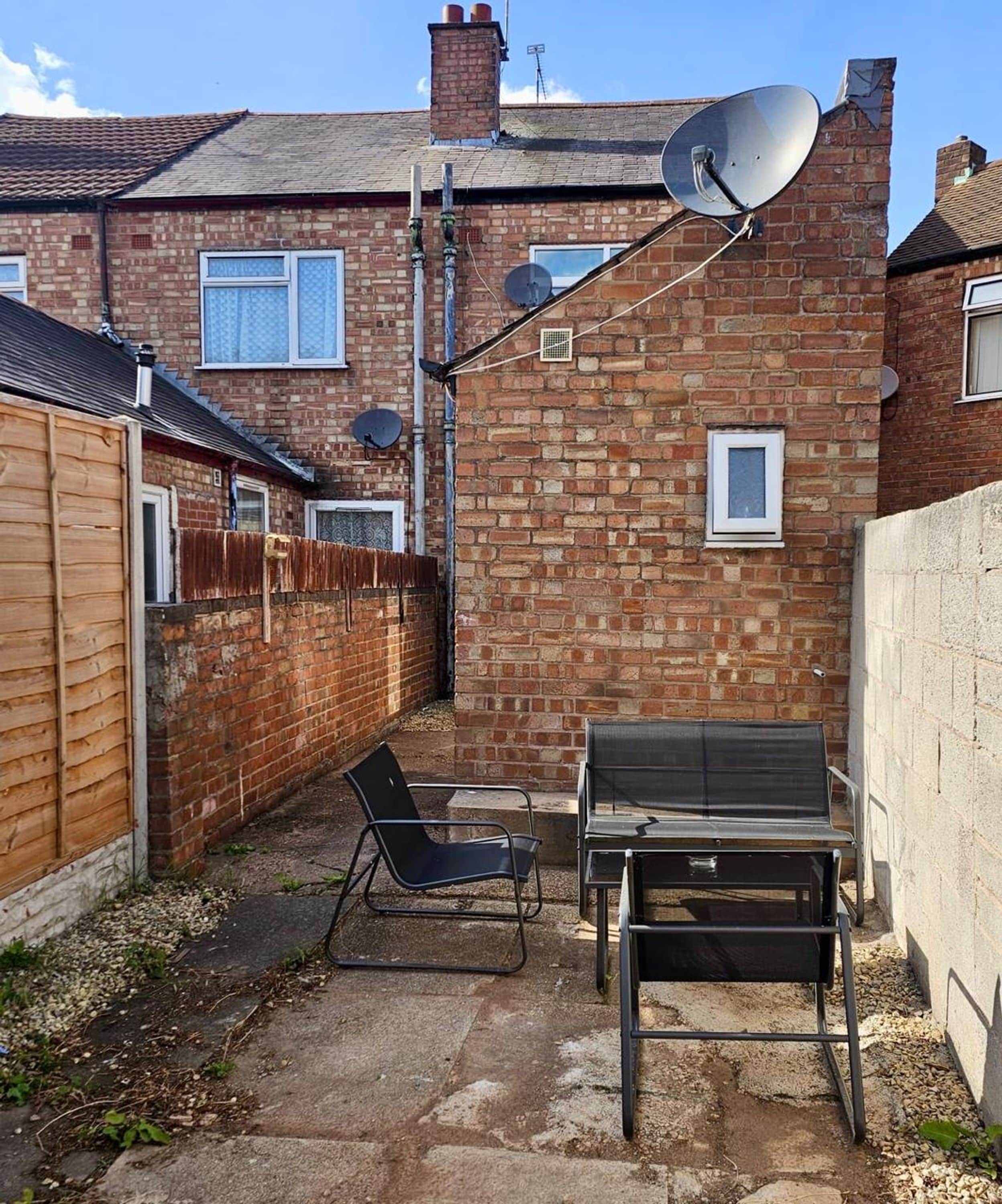A brick house with satellite dishes, a wooden fence, and a patio with two chairs and a table.
