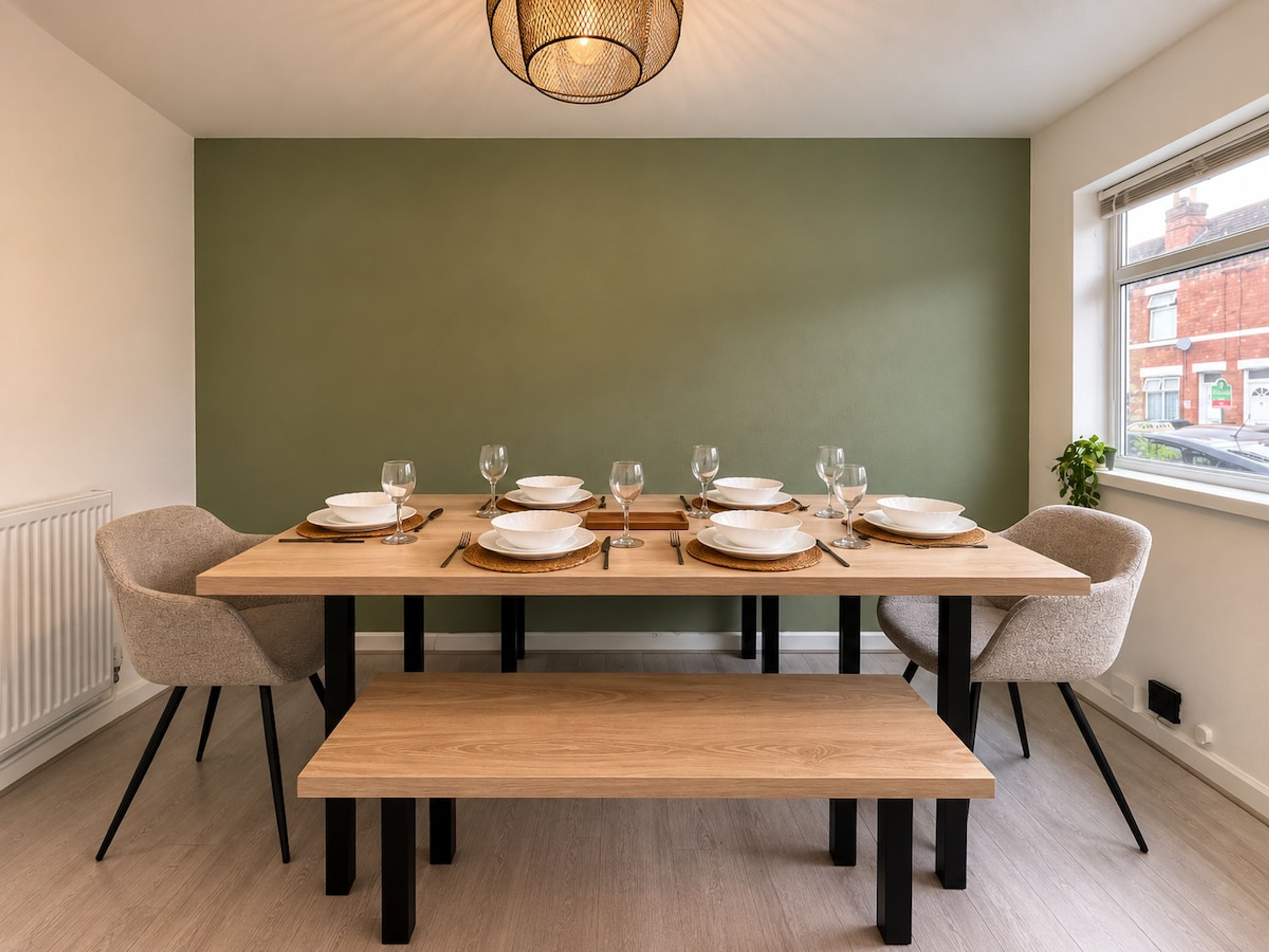 Uma área de refeições com mesa de madeira posta para seis pessoas, com duas cadeiras, um banco e uma parede verde decorativa.