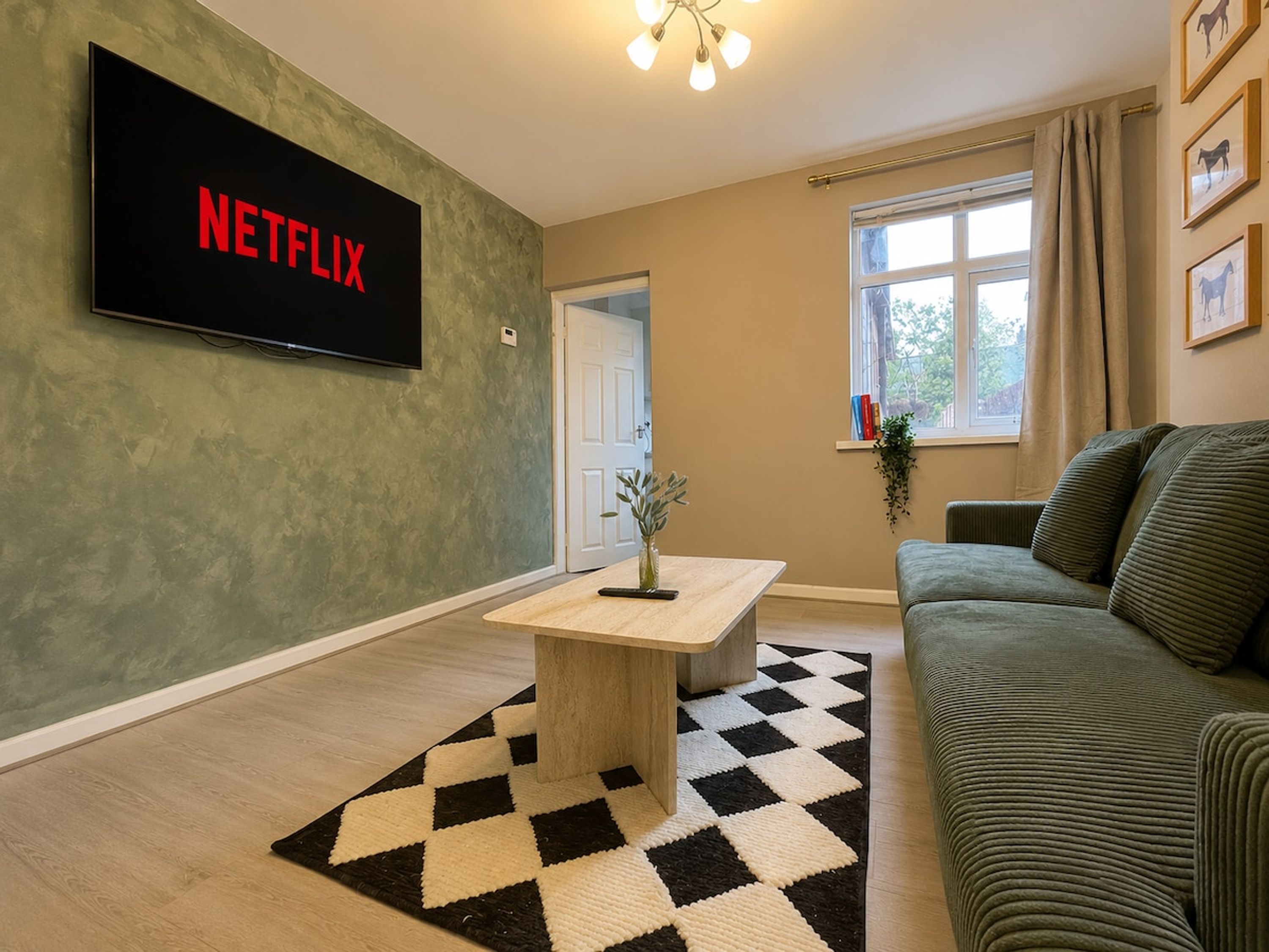 A living room with a flat-screen TV mounted on the wall, a coffee table, a sofa, a window with curtains, and a checkered rug.