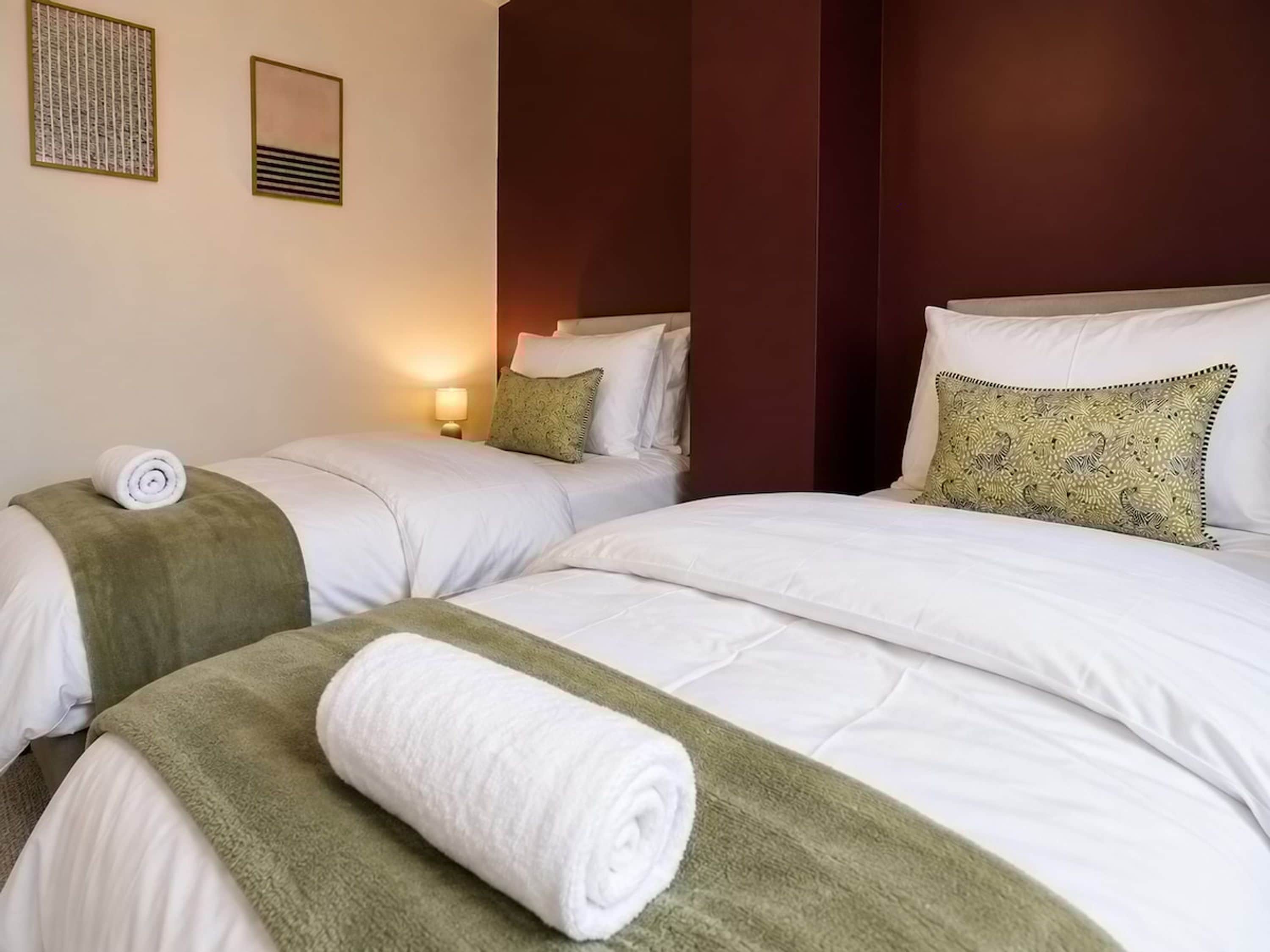 A hotel room with two beds, each with a rolled white towel, a green throw blanket, and a patterned pillow.