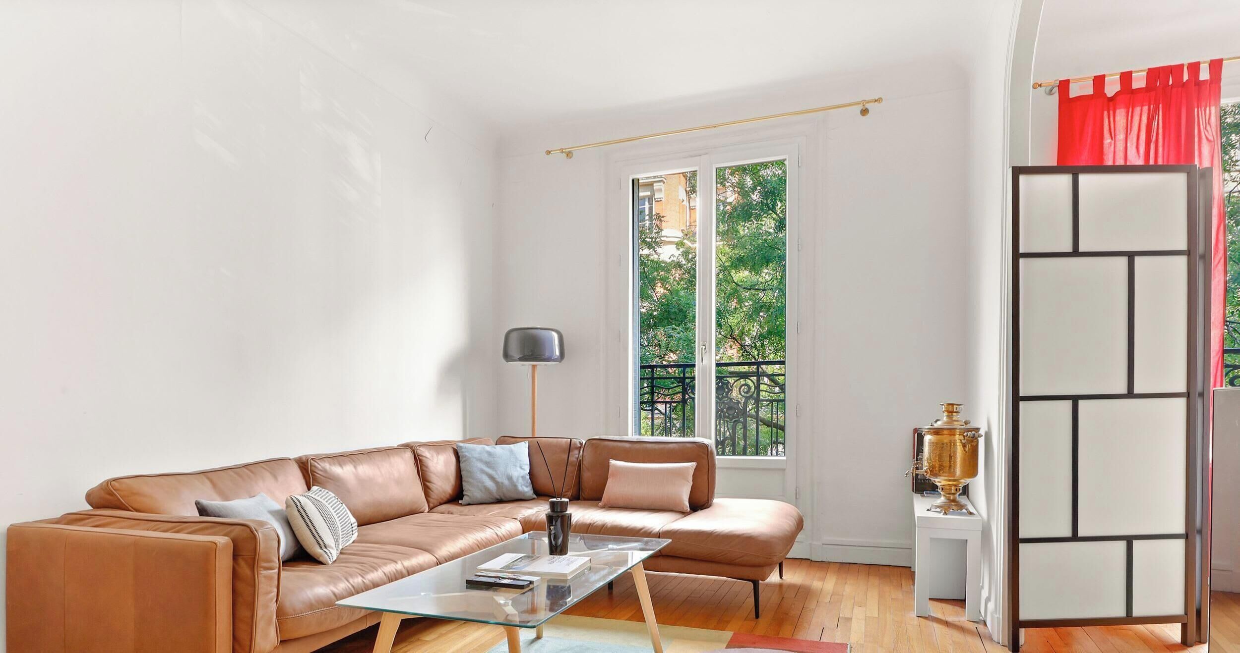A modern living room with a brown leather sofa, a glass coffee table, and a view of greenery through a large window.