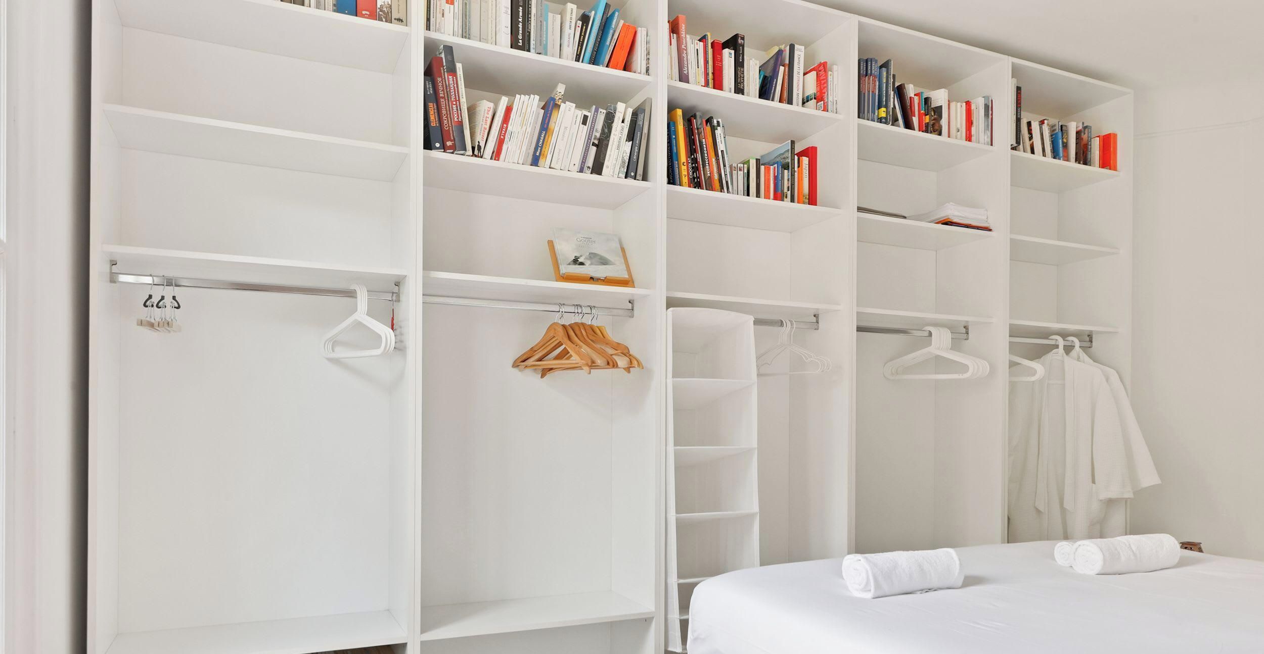 A bedroom with a large white wardrobe, a bed with white linens, and a shelf with books.