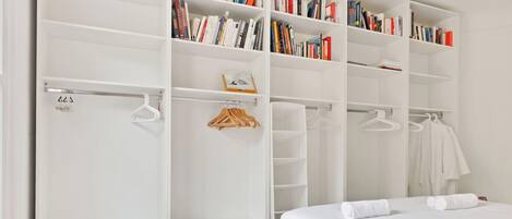 Un dormitorio con un amplio armario blanco, una cama con ropa de cama blanca y una estantería con libros.