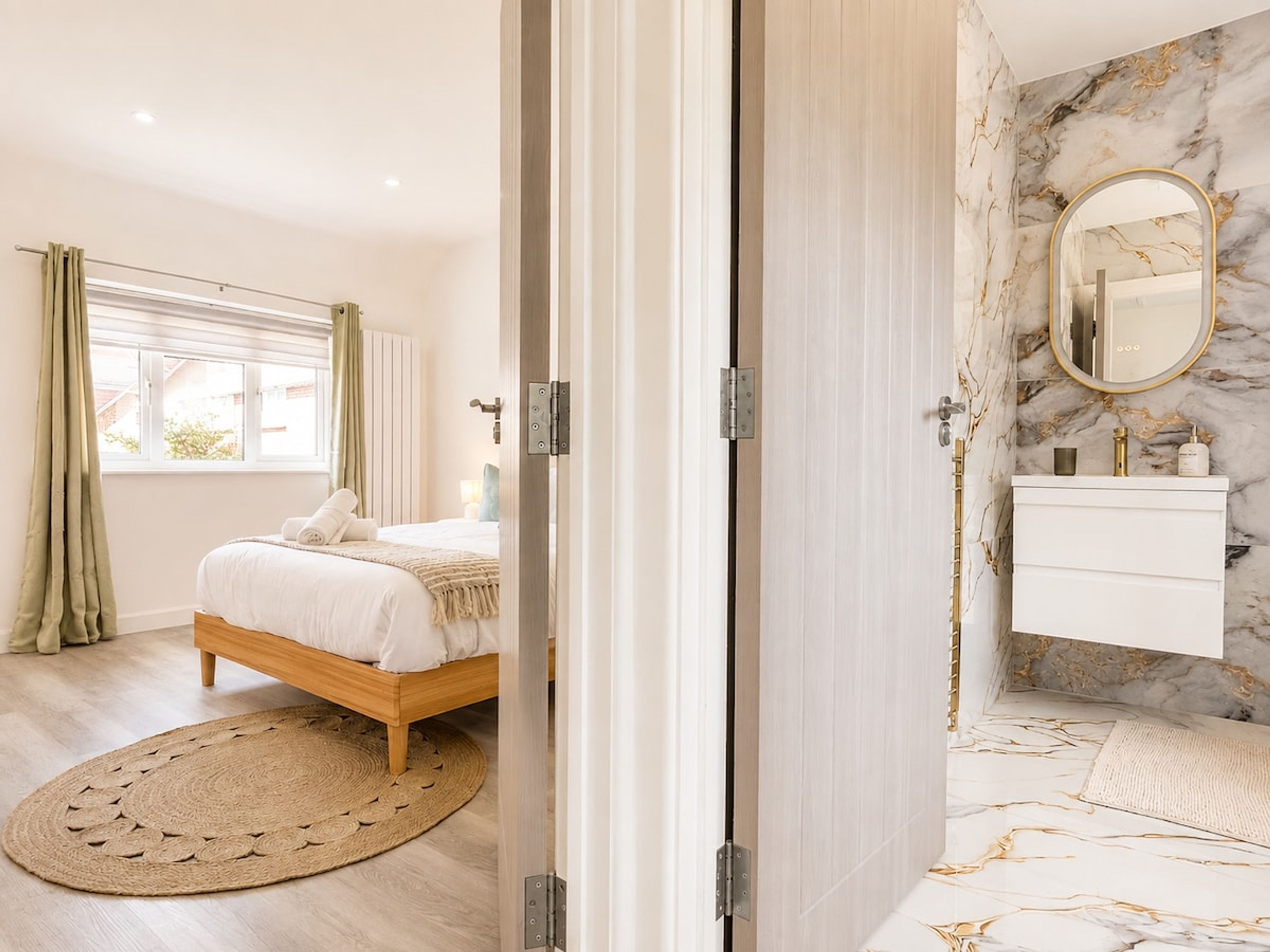 A bedroom with a bed, a round woven rug, a window with curtains, and a bathroom with a marble wall and a mirror.
