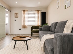 Ein modernes Wohnzimmer mit einem grauen Sofa, einem runden Couchtisch und einem grünen Sessel. Es gibt ein Fenster mit transparenten Vorhängen, eine Heizung und eine Pflanze auf der Fensterbank.