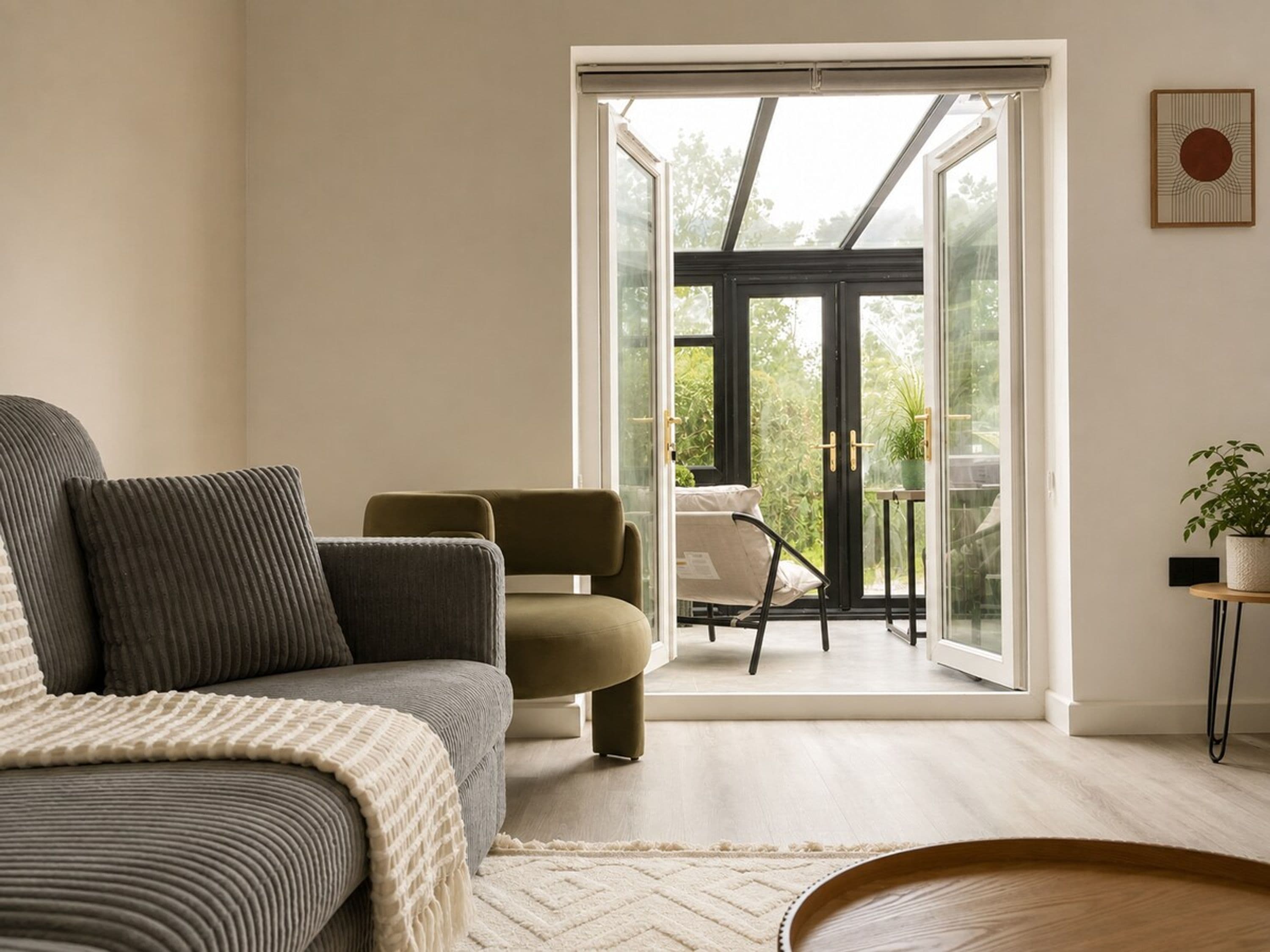 Un salon moderne avec un canapé, un fauteuil et une vue sur un balcon aménagé avec du mobilier d’extérieur.