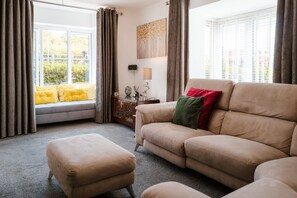 A living room with a sofa, ottoman, and a window with curtains. - Perfect Booking Spacious 3-bed Home w Garden (Rochester)