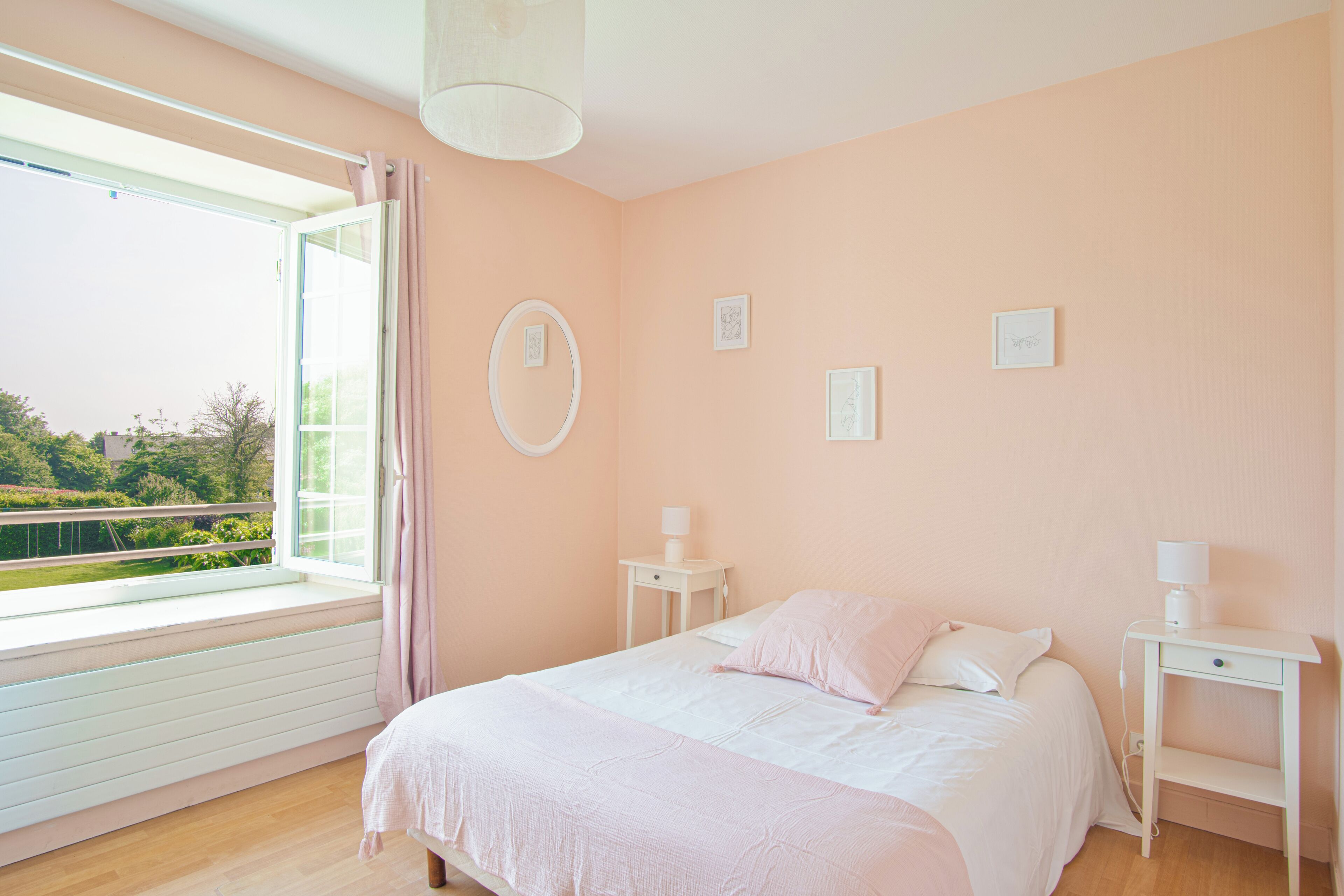 A bedroom with a large window, a bed with a white and pink bedspread, two bedside tables with lamps, and three framed pictures on the wall.