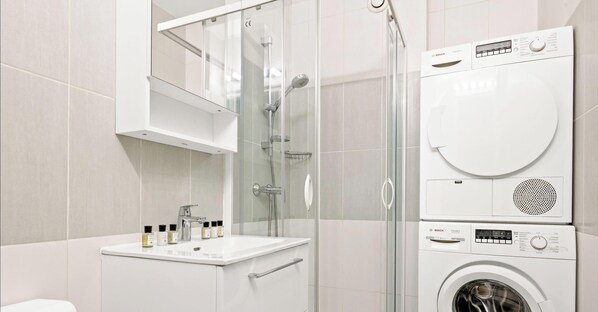 Ein modernes Badezimmer mit Duschkabine aus Glas, Waschbecken mit Pflegeprodukten sowie gestapelter Wasch- und Trockner-Kombination.