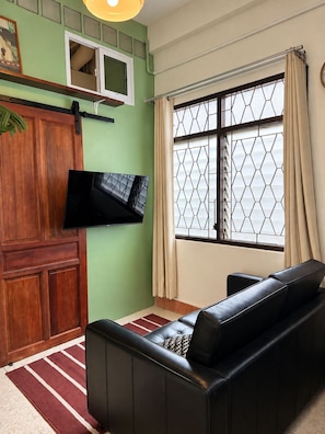 Een woonkamer met een bank, een flatscreen televisie aan de muur, een raam met gordijnen en een houten kast.