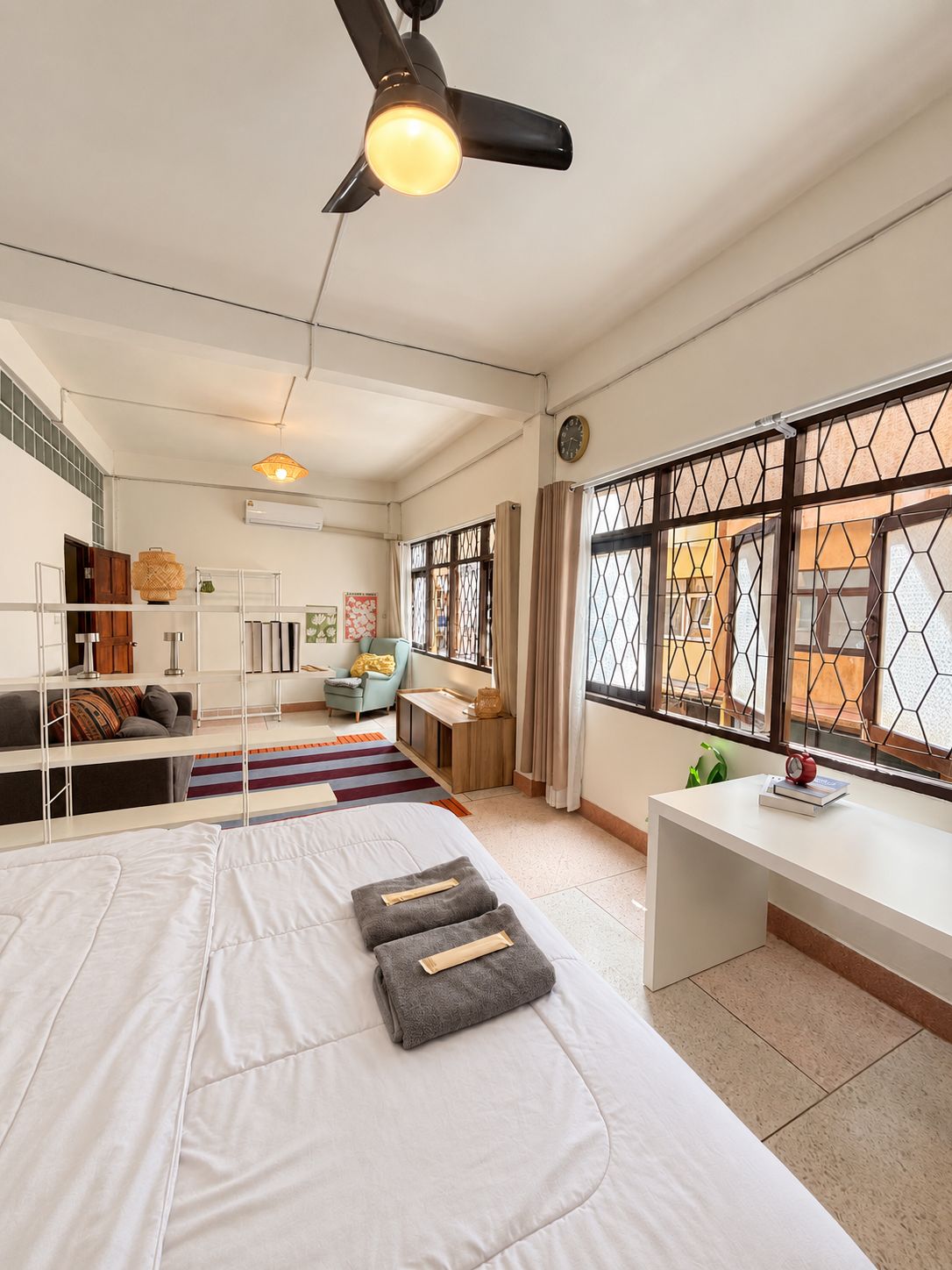 Habitación de hotel con cama, escritorio, silla, ventilador de techo y ventana con celosía decorativa.