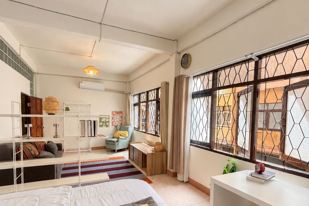 A hotel room with a bed, a desk, a chair, a ceiling fan, and a window with a decorative screen.