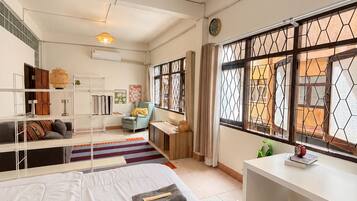 Habitación de hotel con cama, escritorio, silla, ventilador de techo y ventana con celosía decorativa.