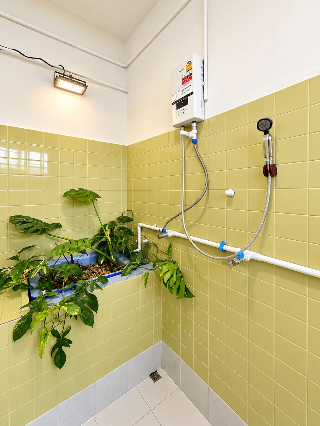 A bathroom with yellow tiles, a shower with a handheld showerhead, a wall-mounted heater, and a plant in a blue planter.