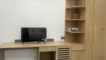 A wooden desk with a flat-screen TV, a power strip, and a shelf with a box and bottles.
