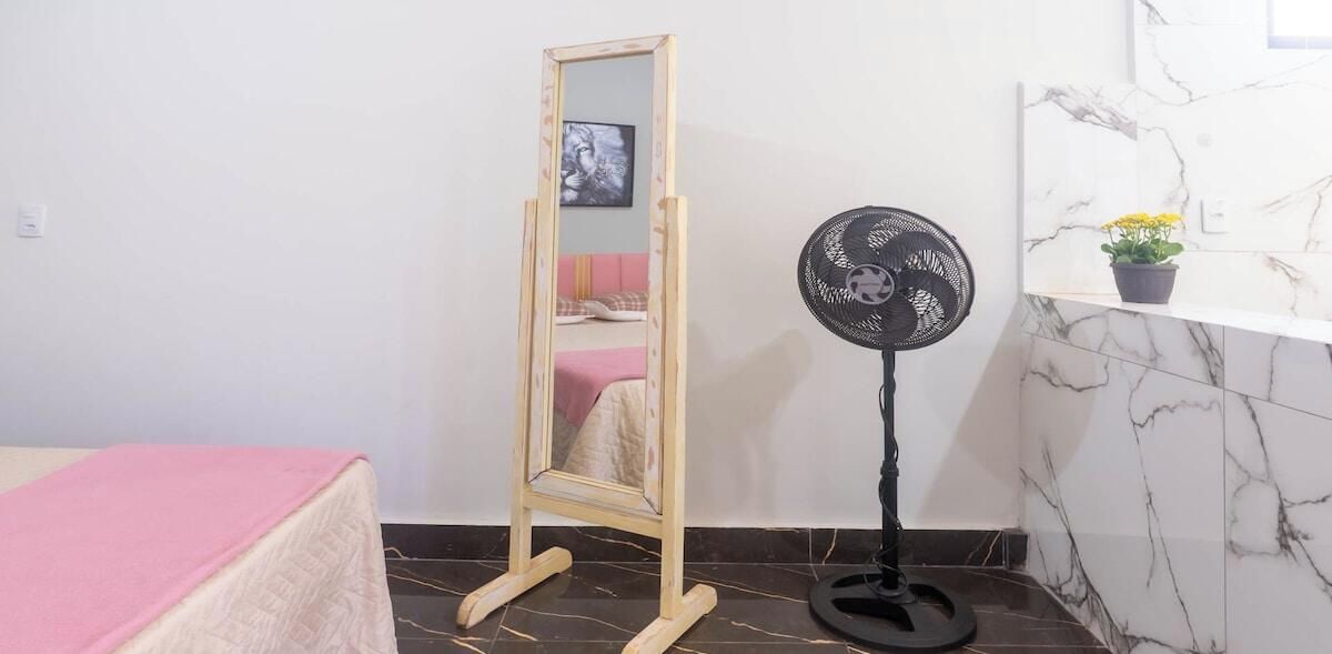 A room with a bed, a standing mirror, a black fan, and a marble-tiled wall.