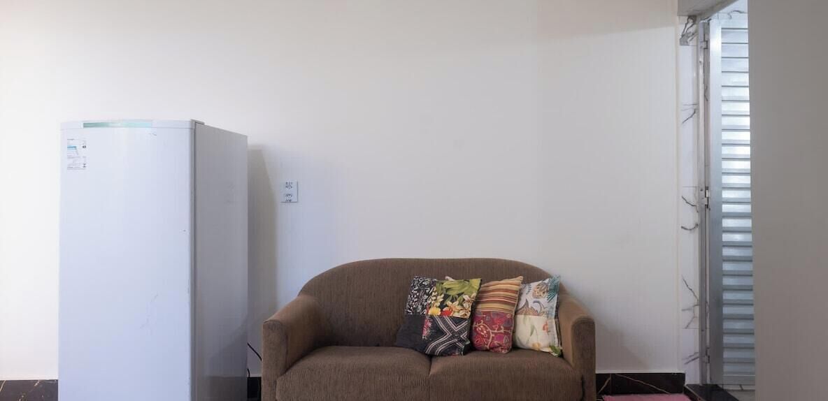 Ein Zimmer mit einem weißen Kühlschrank, einem braunen Sofa mit bunten Kissen und einem Fenster mit Jalousien.