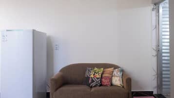Ein Zimmer mit einem weißen Kühlschrank, einem braunen Sofa mit bunten Kissen und einem Fenster mit Jalousien.