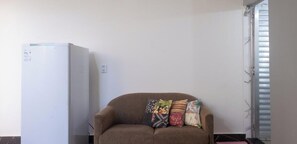 A room with a white refrigerator, a brown sofa with colorful cushions, and a window with blinds.