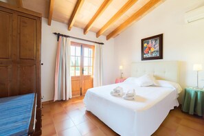 A hotel room with a large bed, a wooden door, a window with curtains, a painting on the wall, and a bedside table with a lamp. - Scenic Country Haven With Pool (Pollença)
