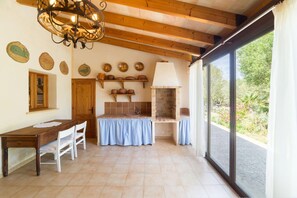 A kitchen with a wooden table, chairs, a hanging light fixture, a tiled backsplash, and a view through large windows. - Scenic Country Haven With Pool (Pollença)