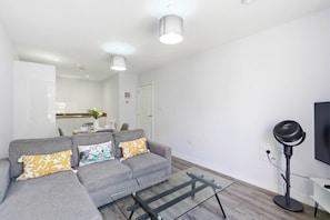 A modern living room with a grey sofa, a glass coffee table, and a standing fan. - Modern Digbeth (Birmingham)