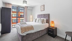 A modern bedroom with a bed, bedside table, and a view of a building through a window. - Modern Digbeth (Birmingham)