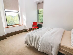 A bedroom with a bed, a red chair, and two windows with views of greenery. - Winnocklands House (Newmilns)