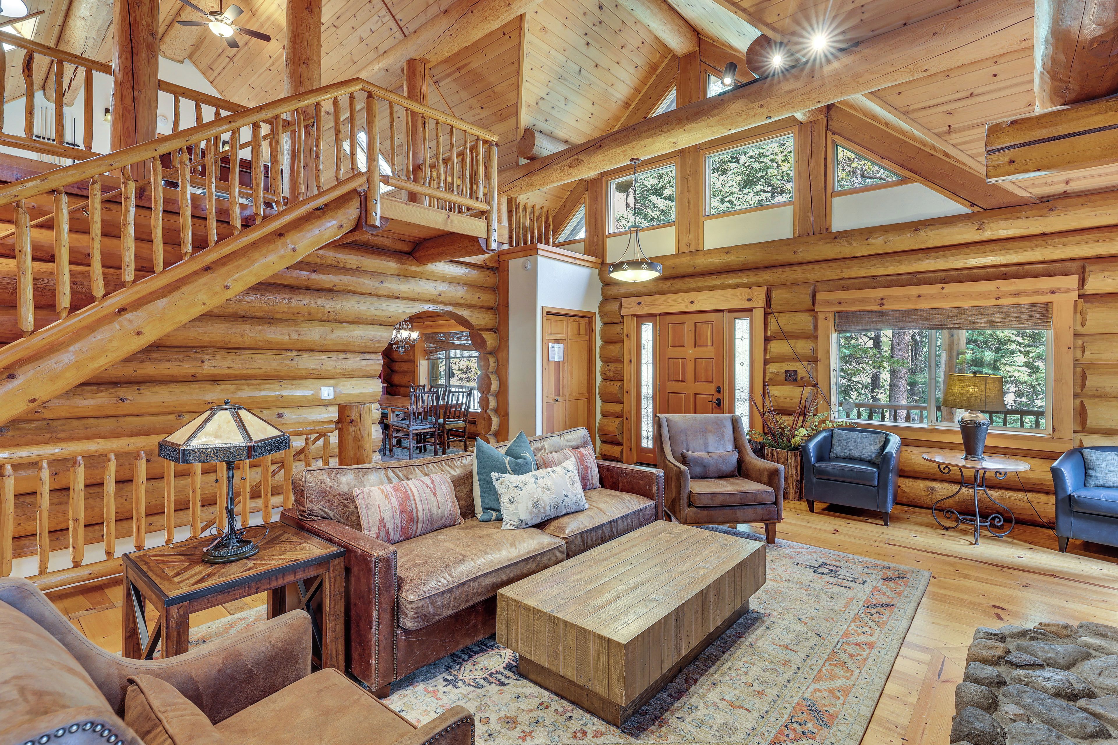A spacious log cabin interior with a wooden staircase, leather furniture, and large windows.