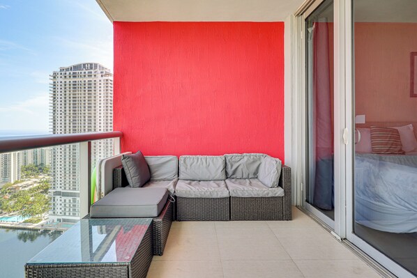 Un balcón con una pared roja, un sofá gris y una mesa de vidrio.