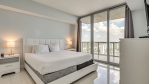 Habitación de hotel moderna con una cama grande, una mesita de noche, una lámpara y vistas a la ciudad a través de un ventanal amplio.