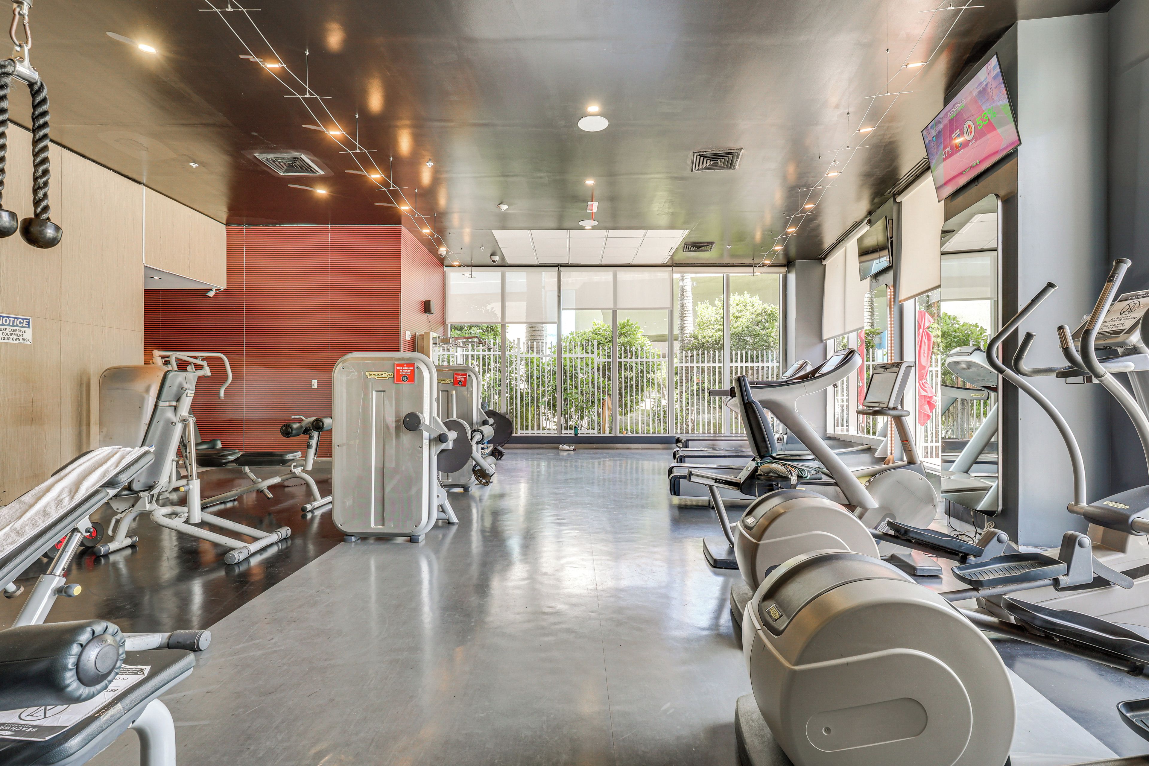 A gym with various exercise equipment, a mirrored wall, and a view of greenery outside.