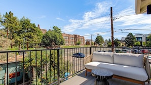 Ein Balkon mit einer Couch und einem kleinen Tisch, mit Blick auf eine Straße mit parkenden Autos und Gebäuden.