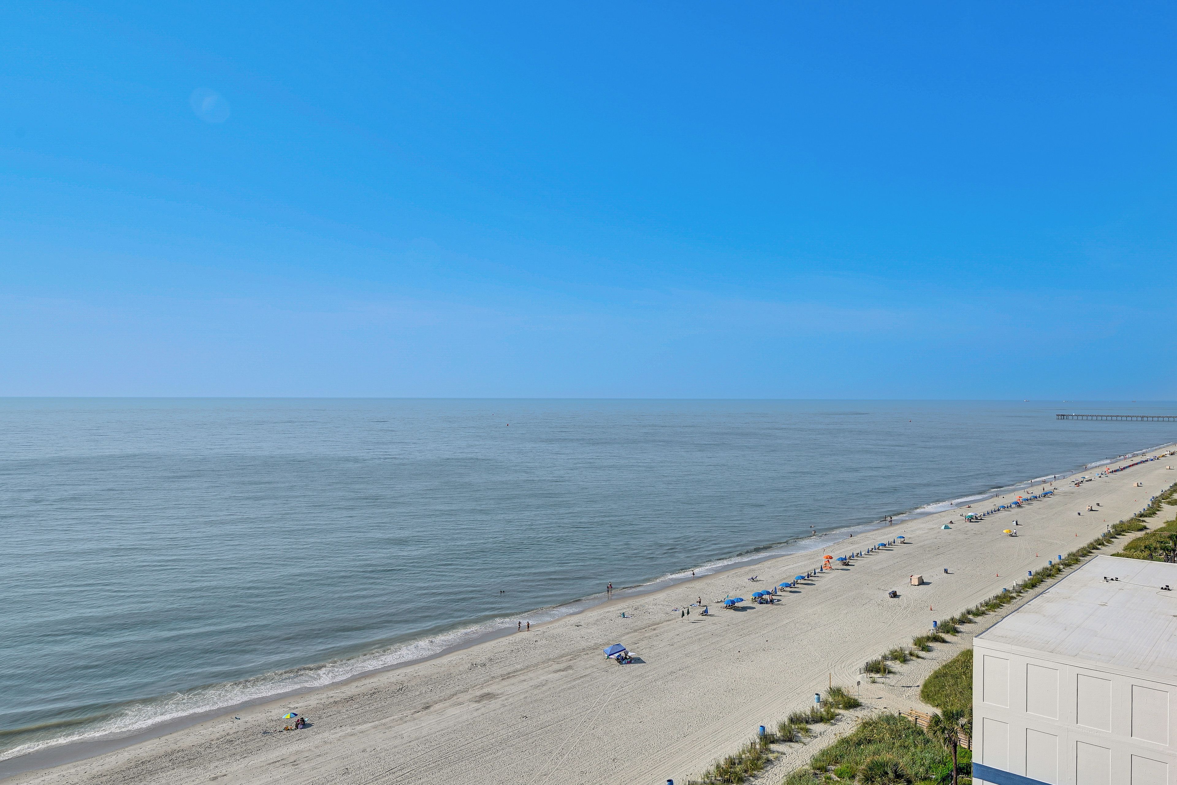A beach with a long stretch of sand, a pier in the distance, and a clear blue sky.