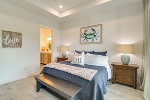 Un dormitorio con cama, mesitas de noche, un banco, una pared con una obra enmarcada y una puerta que da a otra habitación.