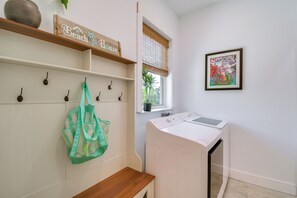 A laundry area with a washing machine, a bench, a wall-mounted bag rack, and a window with a view of greenery. - Fort Morgan Hideaway Hot Tub Golf Beach Access (Gulf Shores)