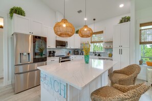 Una cocina moderna con armarios blancos, electrodomésticos de acero inoxidable, una isla de mármol, sillas de mimbre e iluminación colgante.