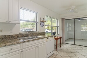 Cocina con armarios blancos, encimera de granito, pileta, reloj y ventana con persianas.