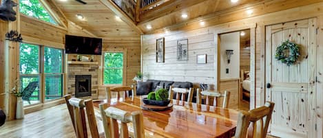 A wooden cabin interior with a dining area, a fireplace, and a staircase.
