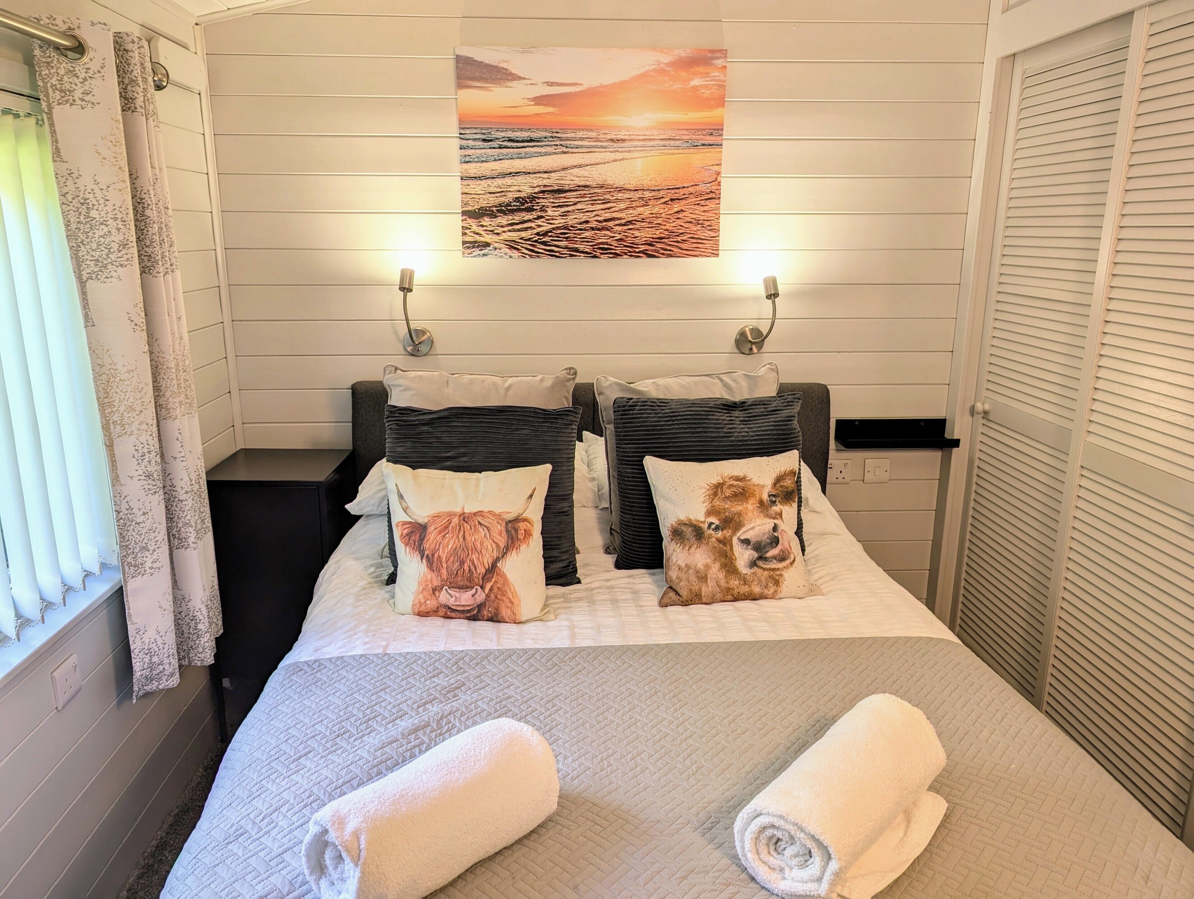 A hotel room with a bed, two pillows with animal prints, a painting above the bed, and a window with curtains.