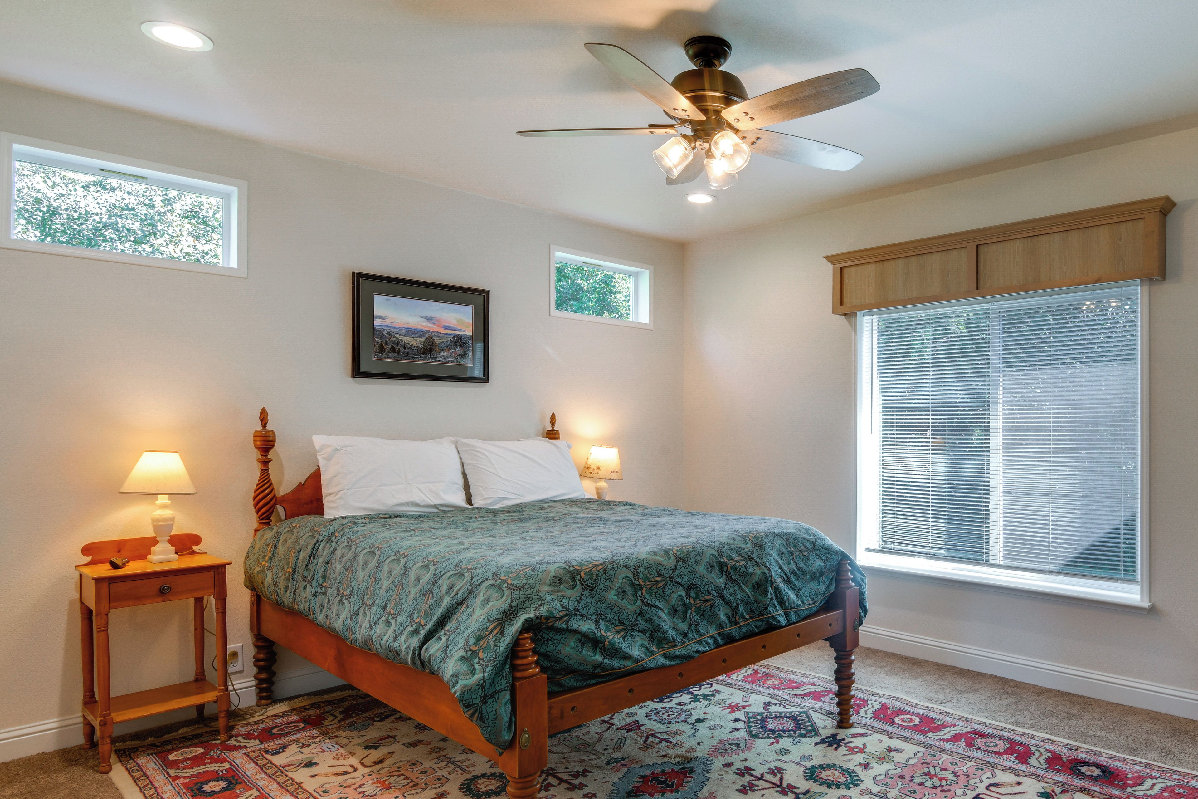 A bedroom with a bed, two bedside lamps, a ceiling fan, a window with blinds, and a rug.