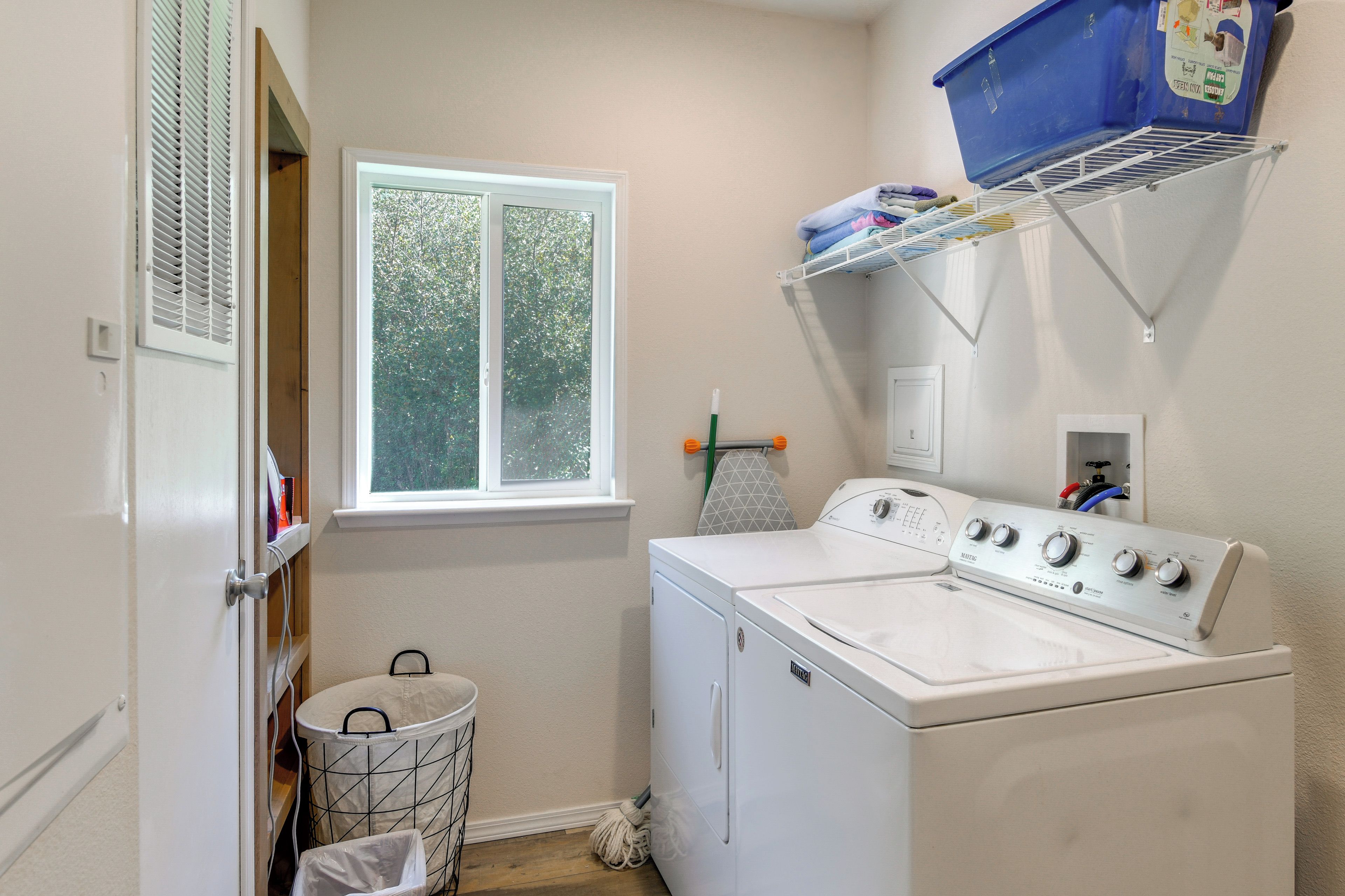 A laundry room with a front-loading washer and dryer, a window, a drying rack, and a trash can.