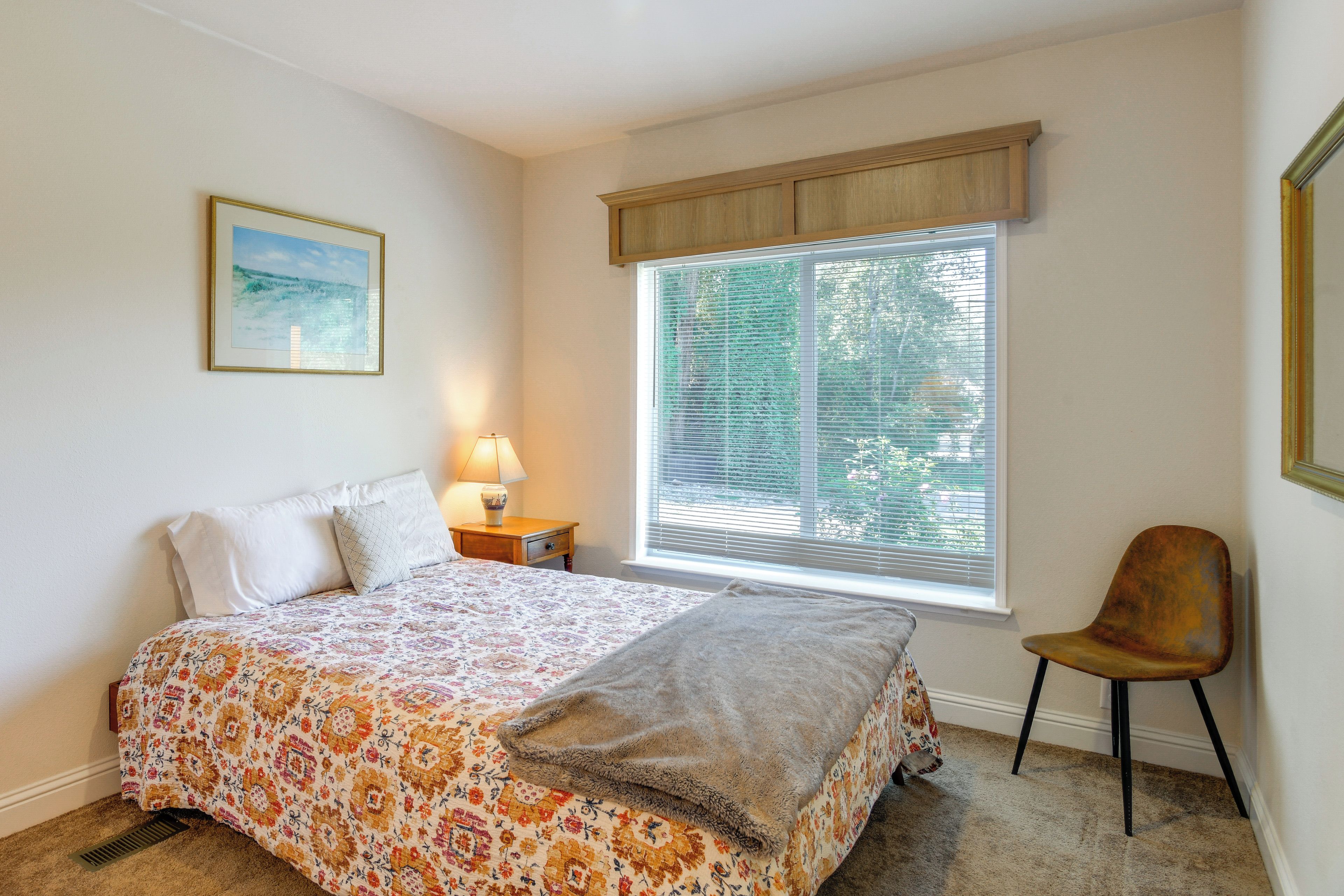 Un dormitorio con cama, una silla, una ventana con persianas, una mesita de noche con lámpara y un cuadro en la pared.