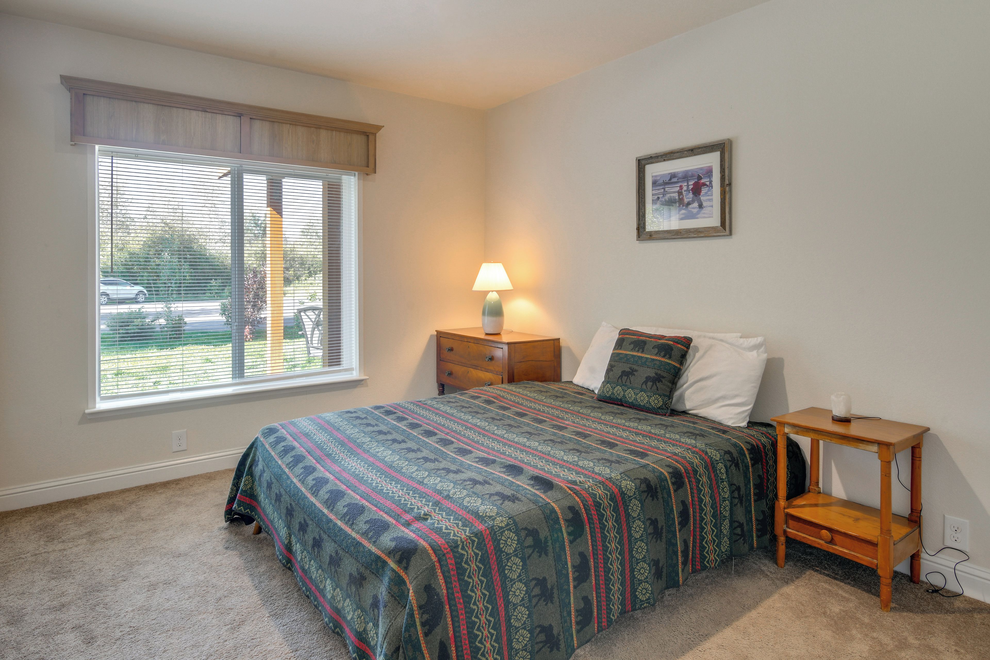 A bedroom with a bed, a nightstand, a lamp, a window with blinds, and a framed picture on the wall.