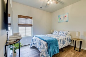 A bedroom with a bed, a bedside table, a lamp, a window with blinds, a ceiling fan, and a painting on the wall.