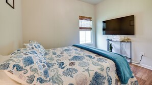 A bedroom with a bed, a window with blinds, a flat-screen TV mounted on the wall, and a small table with towels and a plant.