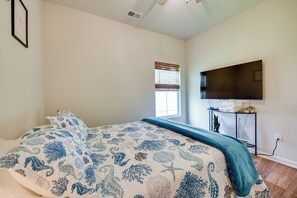 A bedroom with a bed, a window with blinds, a flat-screen TV mounted on the wall, and a small table with towels and a plant.