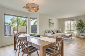 Uma sala de jantar moderna com mesa e cadeiras de madeira, luminária pendente e vista para o exterior por grandes janelas.